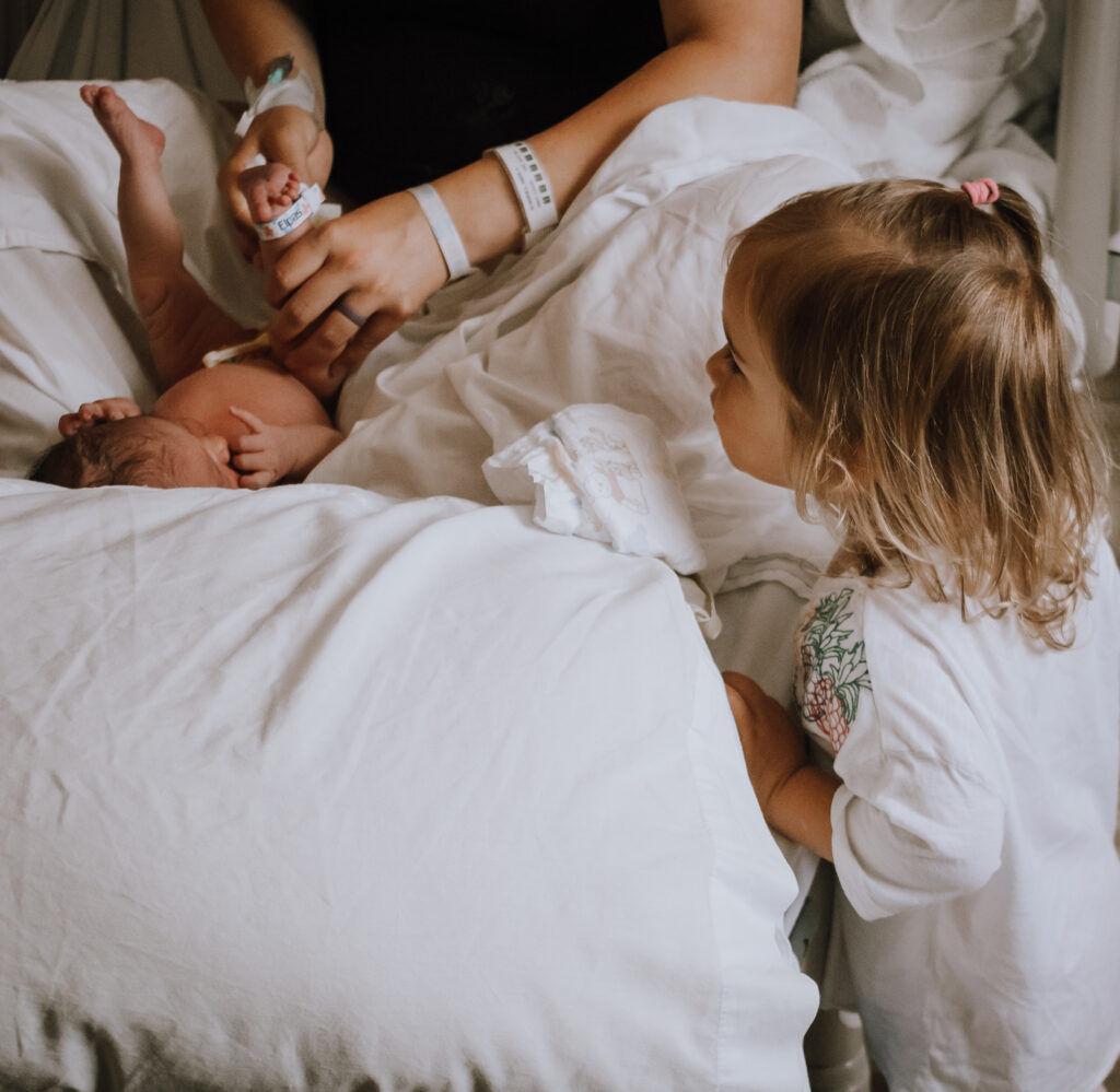Big toddler sister looks over the hospital bed at newborn baby sister while postpartum mom changes baby